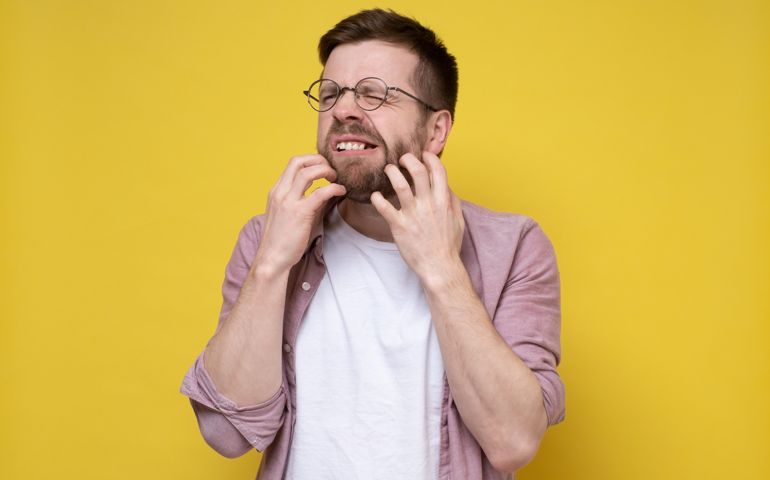 Man in glasses scratches the itchy skin on his chin with hand, he suffers from allergies or irritation from unshaven, bares teeth and closes eyes. Studio, yellow background.