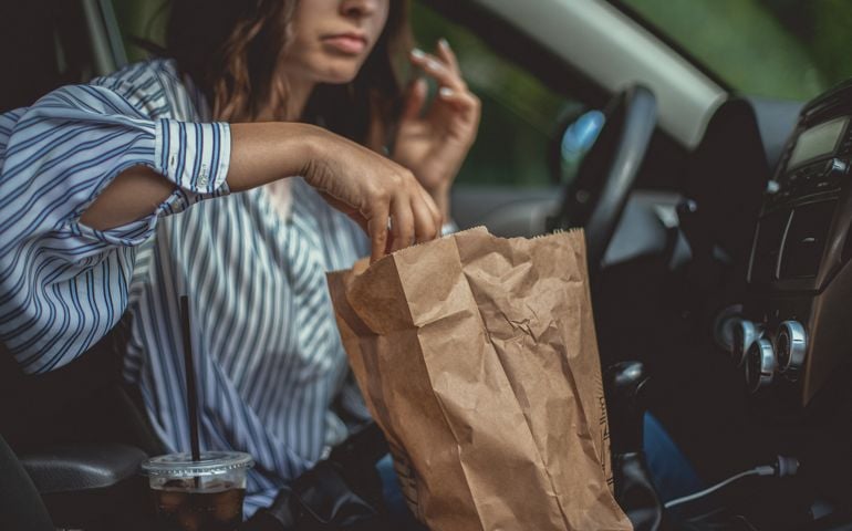 A beautiful young woman is reaching for some french fries in the paper bag and she can't wait to start the brunch in her car.