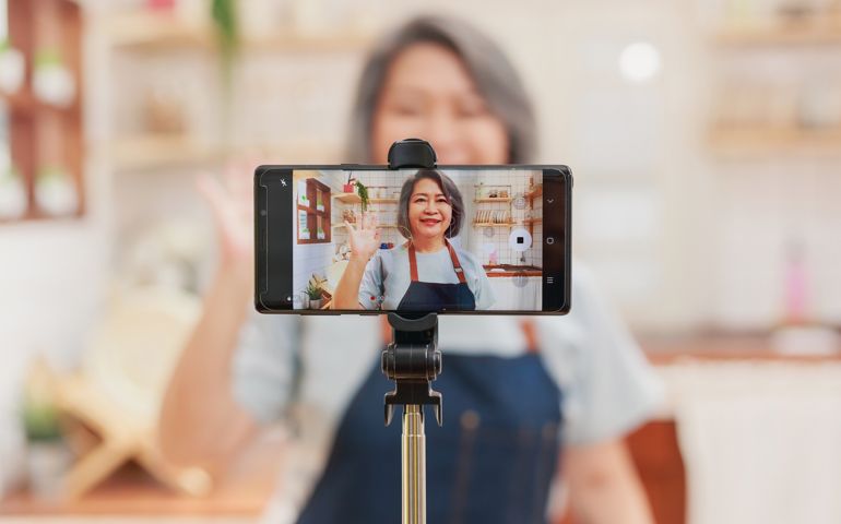 Close up of camera recording pensioner mature woman cooking and making video live for the social media platform. Senior retired influencer woman creating online content video in her kitchen at home.