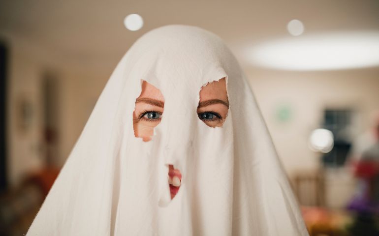 Woman smiling while dressed up as a homemade ghost, her costume consisting of a sheet over her head with eye and mouth holes cut out.