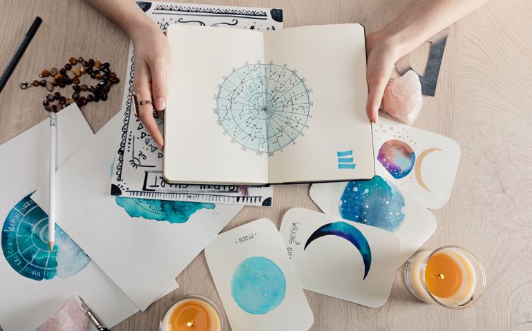 Top view of astrologer holding notebook with watercolor drawings and zodiac signs on cards on table