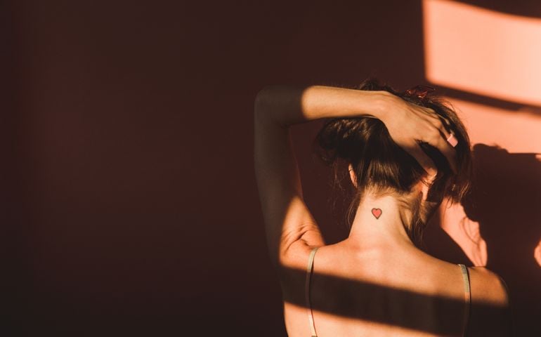 Love, Romance vs. Lonely Isolation: Woman's back and hand through hair, showing heart tattoo. Conceptual image for relationships, self love, and dating. Rear view woman with copy space.