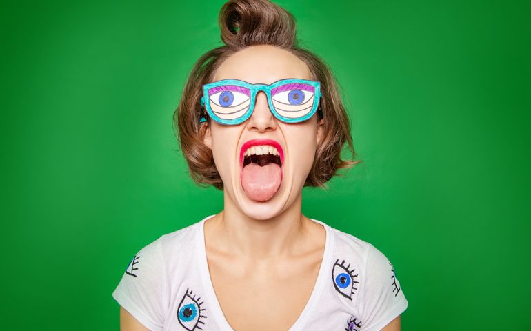 Eccentric young female with funny fake eyewear showing tongue against green background in studio