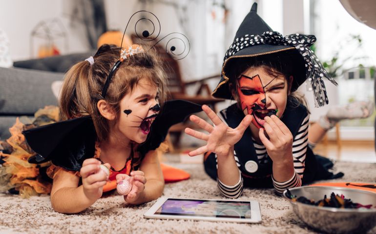 Portrait of the two cute girls wearing Halloween costumes, using tablet and eating candies at home.