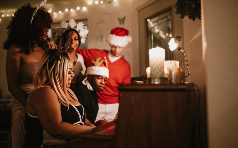A group of young adult friends gather at a home for Christmas celebration over the holiday, dressed to fit the occasion with various Christmas accessories. They sing songs together at the piano, enjoying the Christmas cheer.