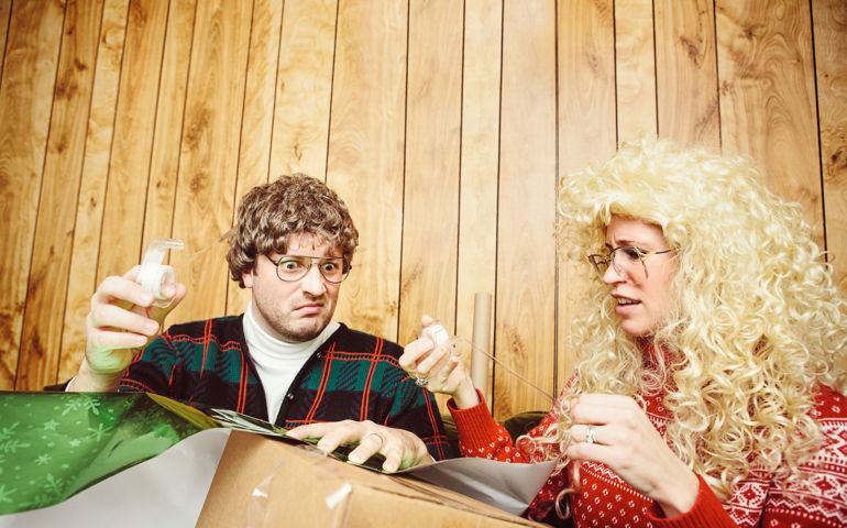 A young married couple in ugly Christmas sweaters feel frustrated and confused as they struggle to wrap a present in gift wrap. Tape and wrapping paper flow freely and make a mess of the room. A Christmas wreath hangs on the wood paneled wall behind them. Horizontal with copy space.
