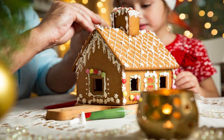 Father and adorable daughter in red hat building gingerbread house together. Beautiful decorated room with lights and Christmas tree, table with candles and lanterns. Happy family celebrating holiday.