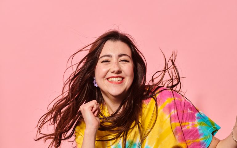 Close up of happy young woman with profound deafness in colourful tie-dye top against pink background laughing and dancing