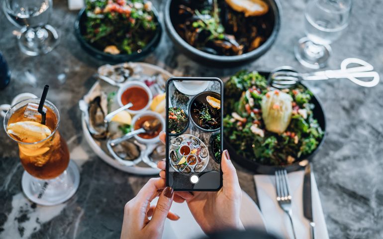 Overhead view of young woman taking photos of scrumptious and delicious meal on dining table with smartphone before eating it in restaurant. Eating out lifestyle. Camera eats first culture