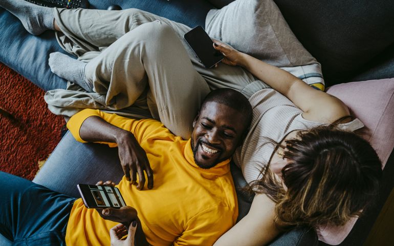 Cheerful man lying on sofa with girlfriend sitting in living room at home