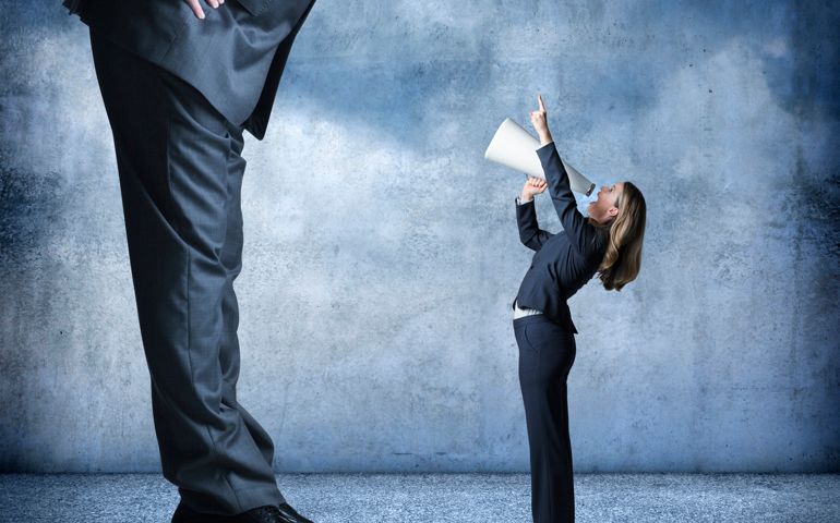 A businesswoman shouts through a megaphone and points up towards a much larger businessman who is standing over her.