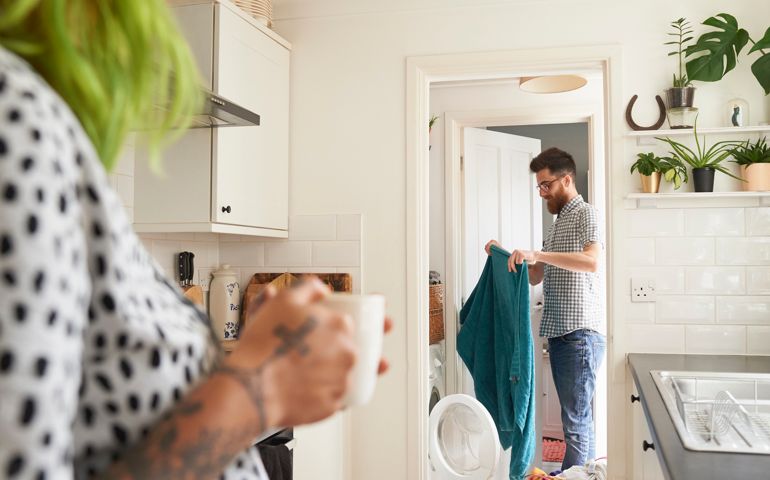 The man is loading the washing machine with laundry while the woman is watching and drinking coffee