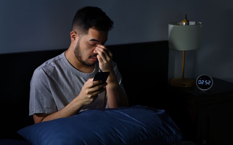 Young man having sore and tired eyes when using smartphone while lying in bed at night