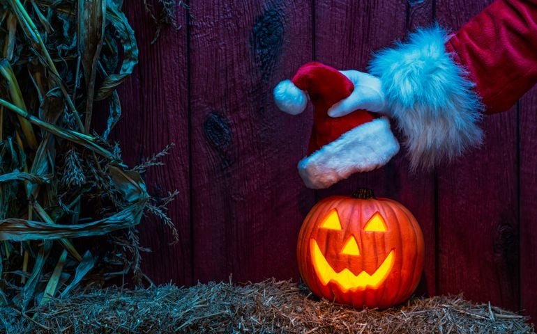 Santa's gloved hand placing a Santa Hat on a carved glowing happy Jack O' Lantern