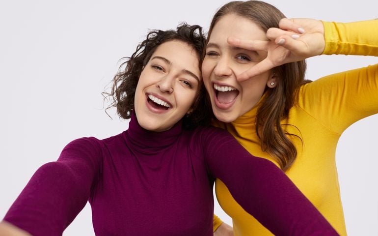 Two best friend smiling and gesturing V sign while taking selfie against white background together