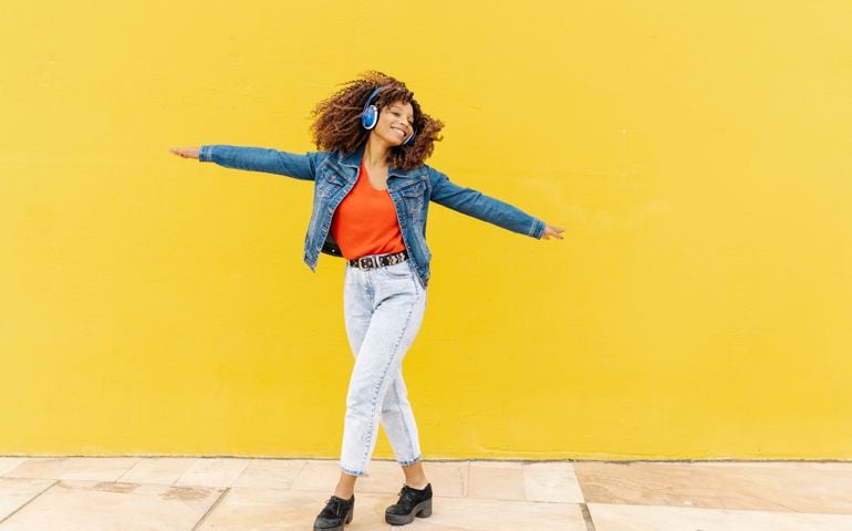 Young woman with curly hair dancing and listening to music with headphones with a yellow wall in the background.