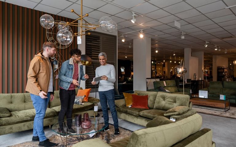 A wide shot of a mid adult couple in a showroom together, they are considering buying new furniture for their home. A store employee is showing them different colour options for a sofa on a digital tablet.