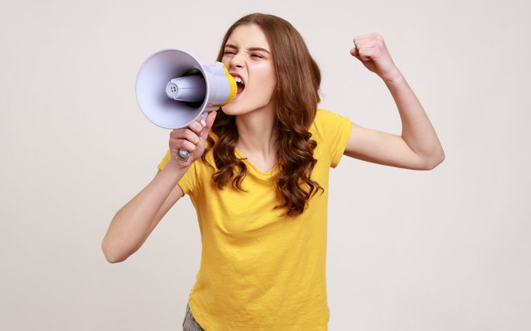 Angry nervous teenager girl with brown hair in yellow t-shirt loudly screaming at megaphone,