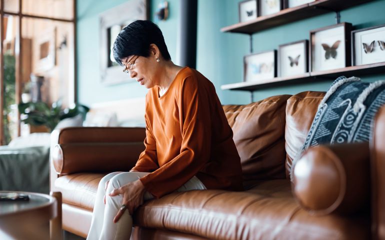 A distraught senior Asian woman feeling unwell, suffering from pain in leg while sitting on sofa in the living room at home