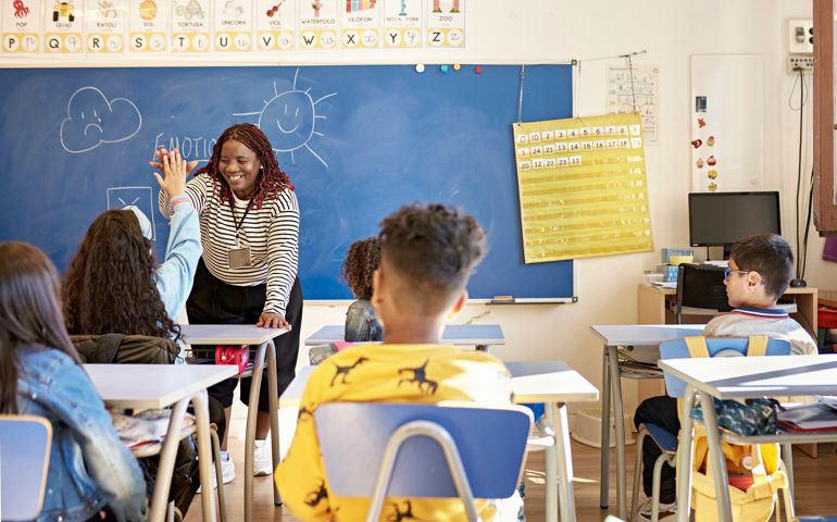 View from behind classmates seated at child-size desks of smiling mid 40s female instructor supporting student with encouraging gesture.