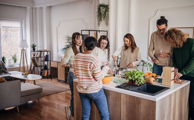 Group of people, diverse male and female friends preparing a meal and drinking wine together in domestic kitchen,