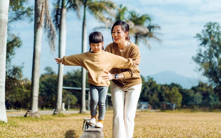 Mother assisting little daughter while balancing on wooden plank with her arms outstretched.
