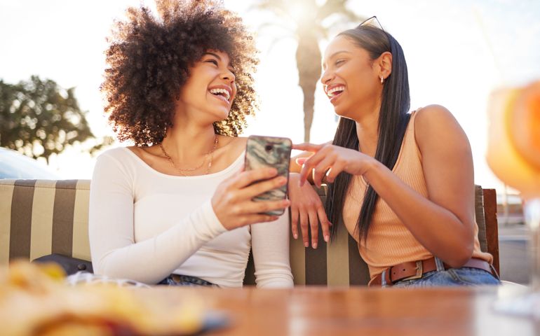 Shot of two friends sharing a funny moment while using a cellphone and having drinks