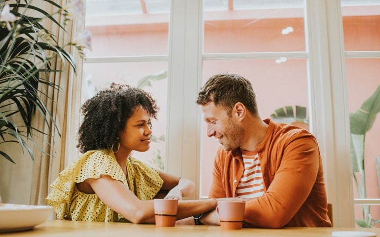 A beautiful young couple looks very much in love as they sit at a dining table sipping hot drinks and flirt.