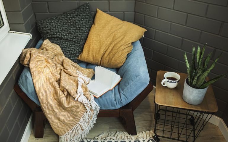 Stylish nook at home for rest and book reading. Veranda decoration. Leisure and relaxation indoors at home. Coffee table with hot drink and house plant. Dark bricks wall.