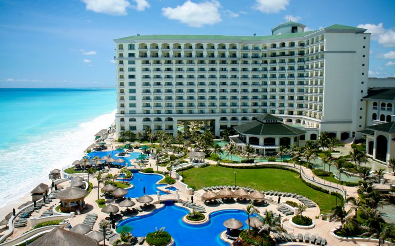 Aerial view of a Cancun resort. There are several large pool areas and a multi-level hotel at this resort. An ocean and sandy beach can be seen to the left of the hotel.