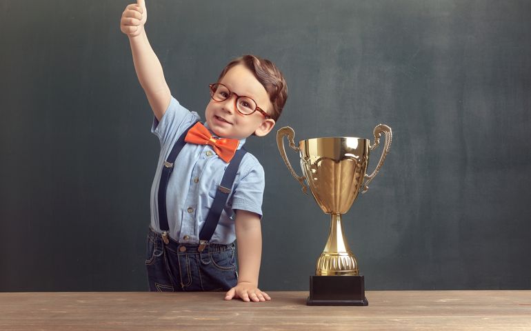 A cute, self-confident 2-3 years old boy is standing and showing his thumb up behind a wooden table with a golden trophy on it. Little boy is wearing an orange bow tie and blue trousers with suspenders.