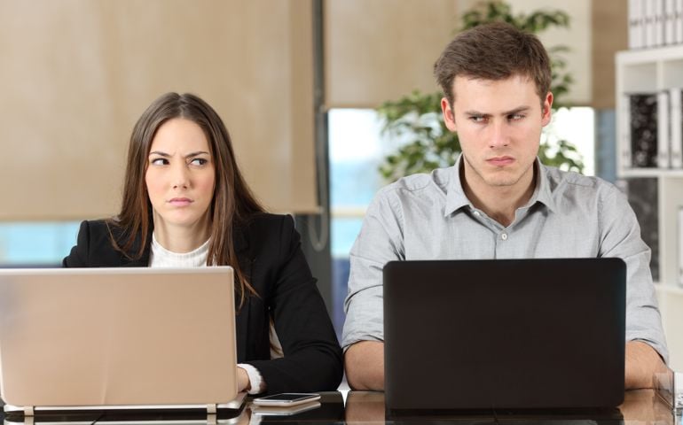Front view of two angry businesspeople using computers disputing at workplace and looking sideways each other with envy