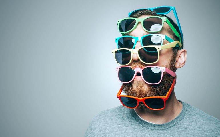 A portrait of a man with a large beard, wearing multiple pairs of multi-colored sunglasses. Studio shot; horizontal with copy space.