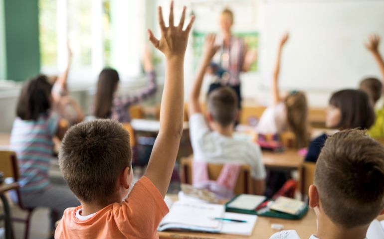 Rear view of little boy and his classmates raising arms to answer teacher's question during the lecture in the classroom.