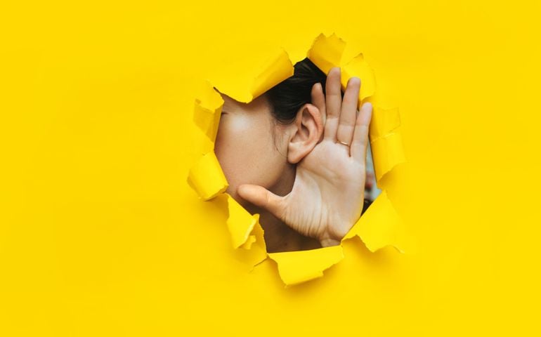 Close-up of a woman's ear and hand through a torn hole in the paper. Yellow background, copy space. The concept of eavesdropping, espionage, gossip and tabloids.