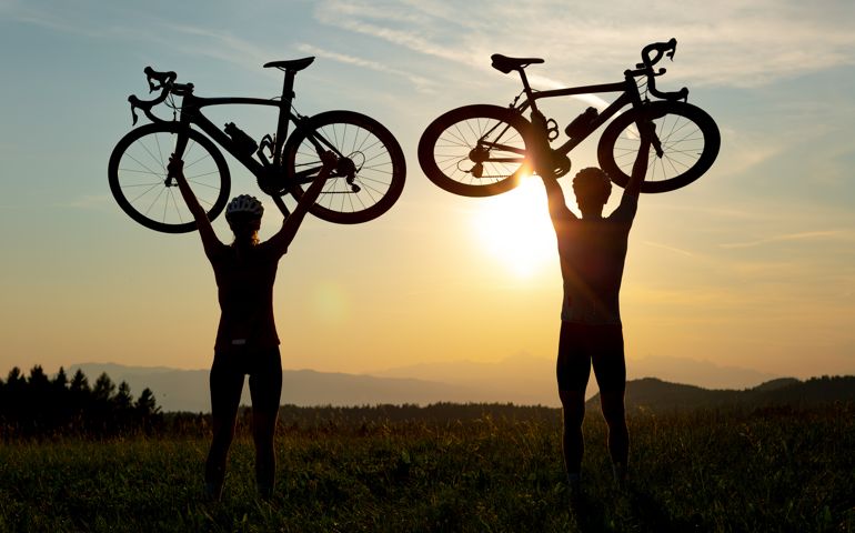 Two silhouettes holding bicycles high above heads, in winner pose
