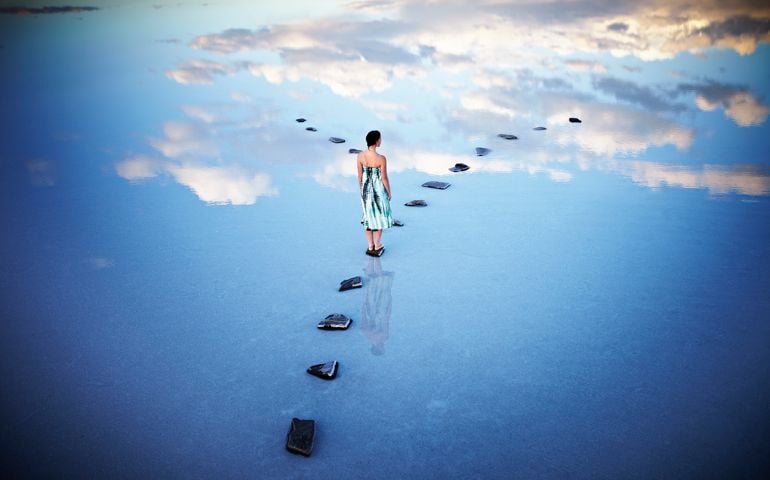 Woman standing at fork in stone pathway in calm lake with clouds reflecting in water