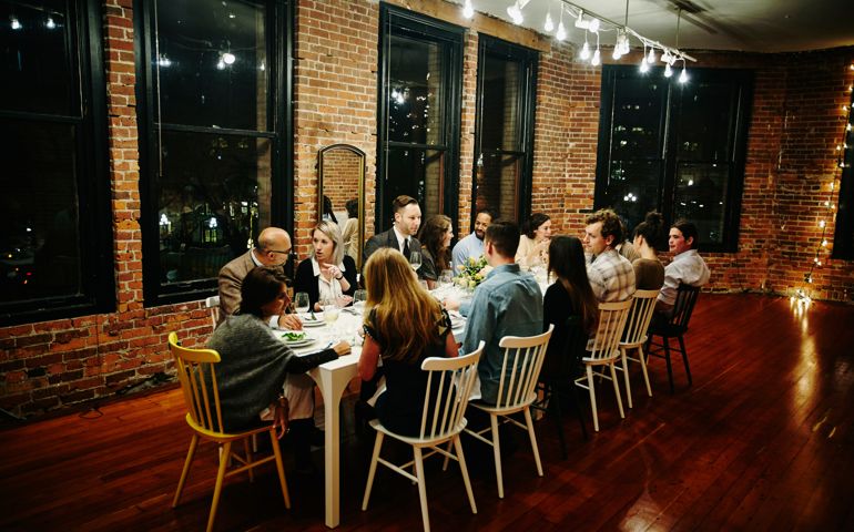 Friends and family sitting gathered together at table during dinner party in loft