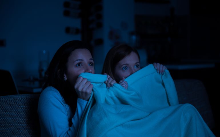 Two women watching horror movie on a tv together, sitting on a sofa with blanket, scared,