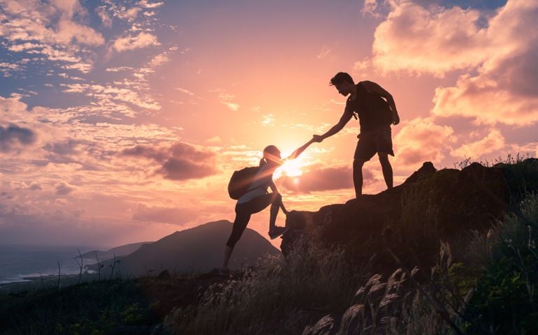 Male and female hikers climbing up mountain cliff and one of them giving helping hand.