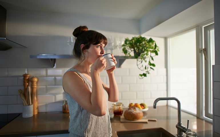 A woman standing in her kitchen drinks from a mug.