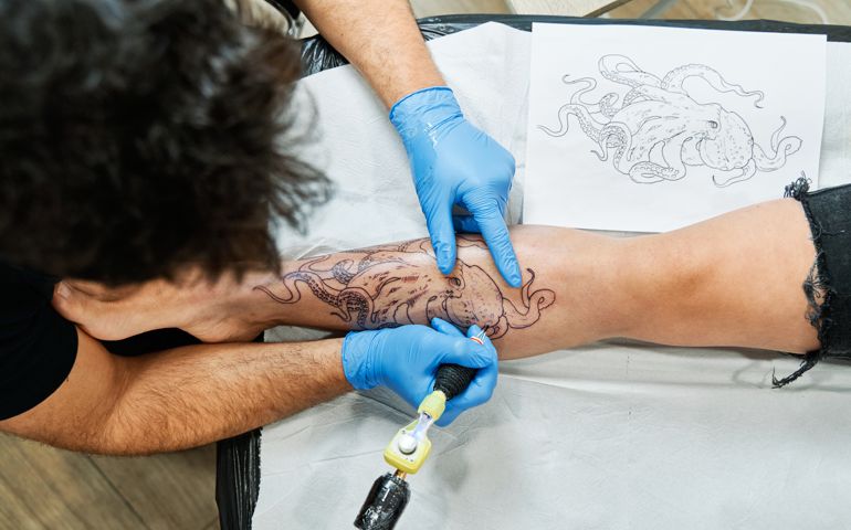 Top view of an unrecognizable tattoo artist tattooing an octopus design on his client's leg.
