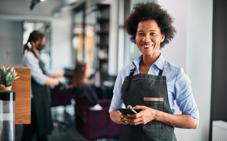 Happy American hair salon owner text messaging on smart phone at work.