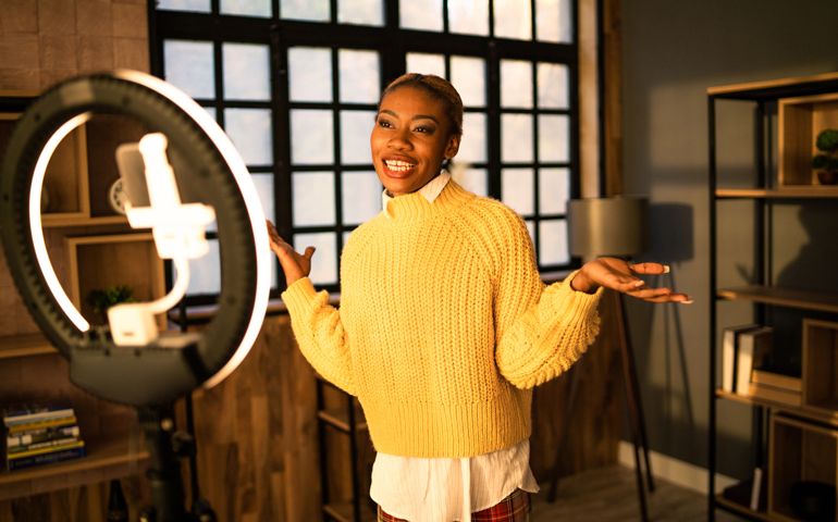 An woman is gesturing with her hands as she films her daily webcast on her cellphone attached to a ring light