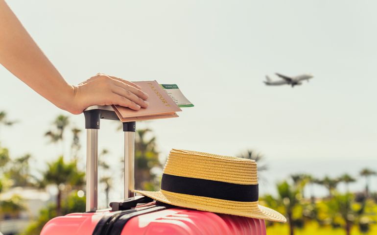 Woman with pink suitcase and passport with boarding pass standing on passengers ladder of airplane opposite sea with palm trees.