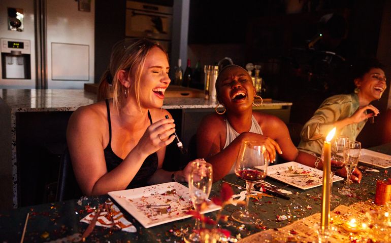 Two diverse young female friends laughing together while sitting at a table covered in confetti during a New Year's Eve dinner party
