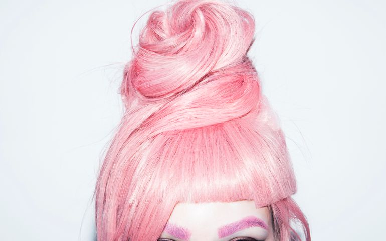 Young woman in her late teens with light blue eyes and pink eyebrows in front of a white background wearing a pink hair wig in an updo
