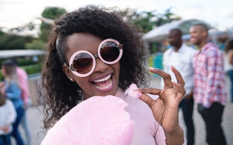 Portrait of a happy woman eating a cotton candy at an amusement park and wearing fun glasses - lifestyle concepts