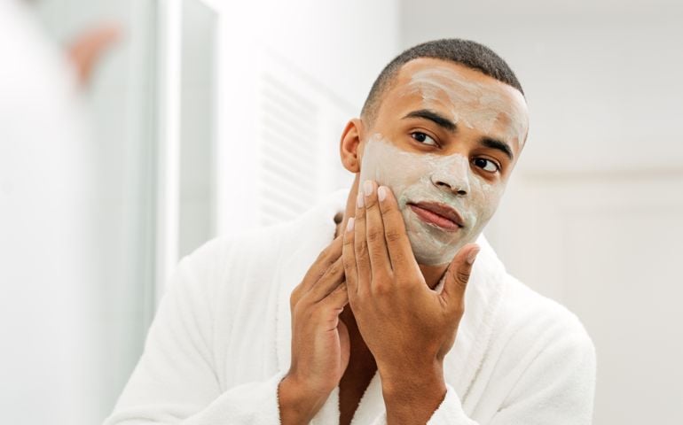 Young Man With Facial Mask Looking In Mirror At Home