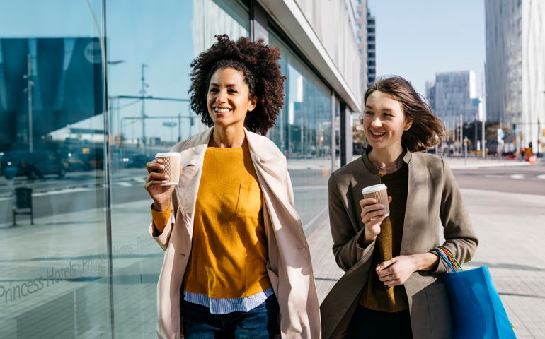 Two happy women with shopping bags and takeaway coffee walking in the city